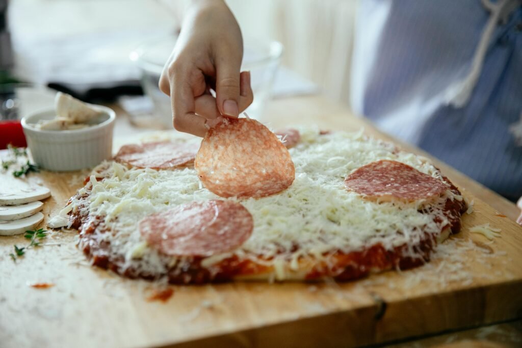 Crop anonymous person putting slice of salami on yummy homemade on wooden cutting board while cooking in kitchen